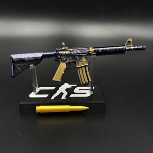 May include: A miniature rifle model featuring a dark blue and gold lightning pattern finish, mounted on a black display stand with the text "CS" and a silhouette figure. A single gold-colored bullet replica rests in front of the display base.