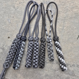 Puede incluir: Un conjunto de siete llaveros de paracord negro, gris y blanco con una variedad de patrones de nudos.