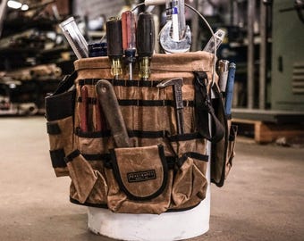 May include: A brown canvas tool bucket with multiple pockets and compartments. The bucket is filled with various tools, including screwdrivers, a wrench, a hammer, and a measuring tape. The bucket is labeled "ReadyMade" on the front.