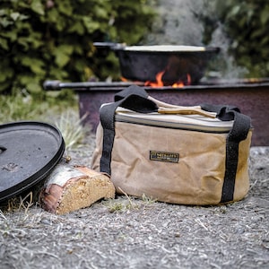 May include: A tan canvas bag with black straps and a zipper closure. The bag has a brown leather handle and a label that reads "Red Owyhee Supply Co.". The bag is sitting on the ground next to a cast iron pot and a piece of firewood.