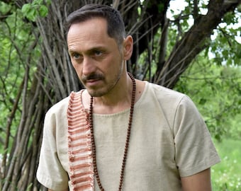 May include: A man wearing a light brown linen shirt with a long, fringed, rust-colored scarf and a brown wooden beaded necklace.