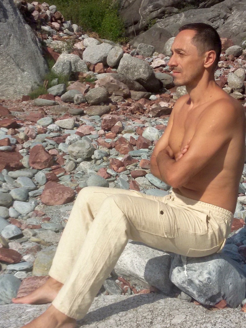 May include: A man wearing beige linen trousers sits on a rock by a rocky shore. The trousers have a large pocket on the right thigh.