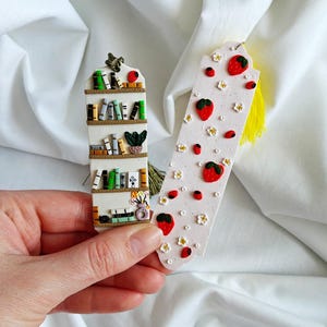 Op de afbeelding: Twee handgemaakte bladwijzers. Eén is gevormd als een boekenplank met miniatuurboeken, een plant en een bloemenvaas. De andere is wit met rode aardbeien, kleine rode stippen en witte bloemen. Beide hebben een kwast.