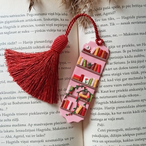 Pode incluir: Um marcador de página rosa com um design de estante de livros. A estante está cheia de livros coloridos e tem uma pequena planta na prateleira superior. O marcador de página está preso a um tassel vermelho.