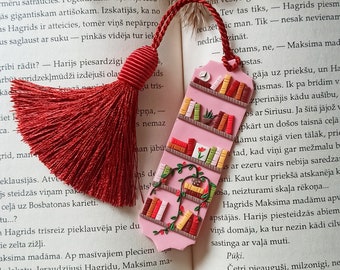 May include: A pink bookmark with a bookshelf design. The bookshelf is filled with colorful books and has a small plant on the top shelf. The bookmark is attached to a red tassel.
