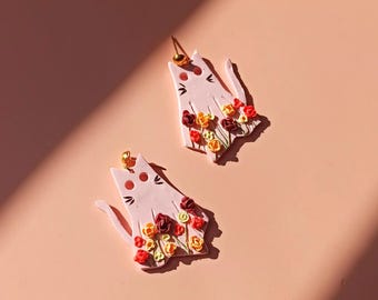 May include: A pair of pink cat-shaped earrings with a floral design. The flowers are red, orange, and yellow.