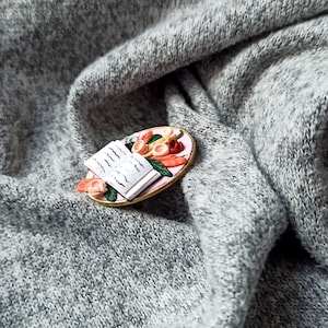 Pode incluir: Broche em miniatura com tema de comida, apresentando um pequeno livro aberto, vegetais e outros alimentos em um prato oval rosa com borda dourada. O broche está sobre um fundo de tecido de malha cinza.