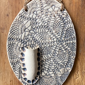 Puede incluir: Colgante de pared de cerámica blanca con un patrón de encaje negro decorativo y un pequeño jarrón adjunto. El colgante tiene un lazo de cuerda para colgar.