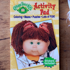May include: A Cabbage Patch Kids activity pad featuring a colourful illustration of a Cabbage Patch Kid doll on the cover. The text on the cover reads "Cabbage Patch Kids Activity Pad Coloring Mazes Puzzles Lots of FUN!" and "Stickers Included".