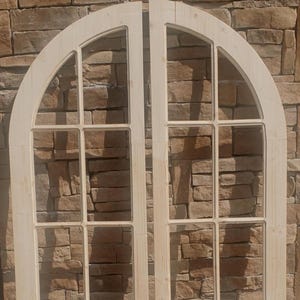 May include: Two unfinished wooden arched doors with multiple rectangular window panes. The doors are made of light-colored wood and are set against a textured brick wall. The doors have a classic, elegant design.