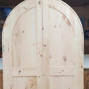 May include: A pair of unfinished wooden doors with an arched top. The doors are made of light-colored wood with visible grain and knots. Each door has a rectangular panel at the bottom and a curved top section.