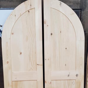 May include: Two unfinished wooden arched doors. Each door is made of light-colored wood with visible grain and knots. The doors have a rectangular panel at the bottom and a curved top, creating an arched shape.
