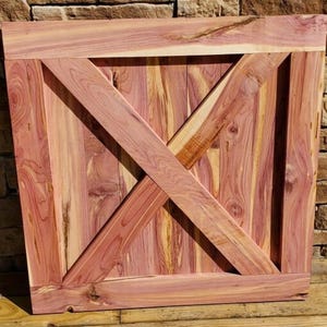 May include: A rectangular wooden panel with a diagonal cross design. The panel is made of reddish-brown wood with visible grain patterns. The cross is formed by two intersecting planks, creating an "X" shape across the panel's surface. The panel is framed by a border.