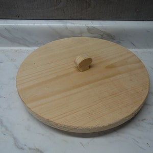 May include: A round wooden lid with a small wooden knob on top. The lid is made of light-colored wood and has a smooth surface.
