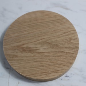 May include: A round, light brown wooden coaster with visible wood grain. The coaster is made of a single piece of wood and has a smooth, finished surface. The coaster is designed to protect surfaces from drink rings.