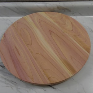 May include: A round wooden cutting board with a natural wood grain pattern. The board is made of light brown wood with reddish and cream-colored streaks. The circular board is approximately 12 inches in diameter.