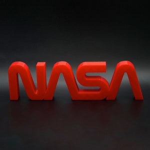 Puede incluir: Letras de plástico rojo en 3D que deletrean "NASA" sobre un fondo negro.