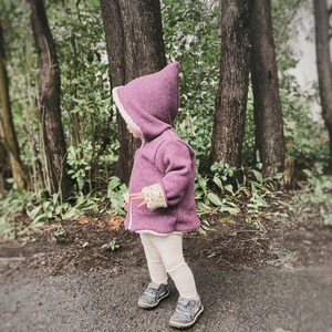 May include: A young child wearing a purple hooded jacket with a white fleece lining and white pants. The jacket has a floral pattern on the cuffs. The child is standing in a wooded area.
