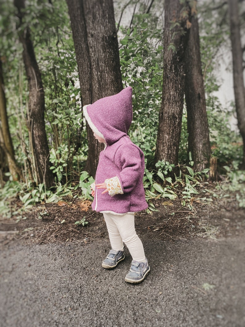 May include: A young child wearing a purple hooded jacket with a white fleece lining and white pants. The jacket has a floral pattern on the cuffs. The child is standing in a wooded area.