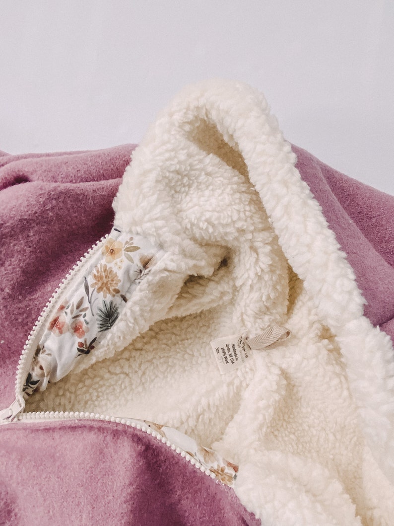 May include: A mauve hooded jacket with a white sherpa lining and a floral print on the inside of the hood. The tag reads "Made in USA, 100% Wool, Size 12M".
