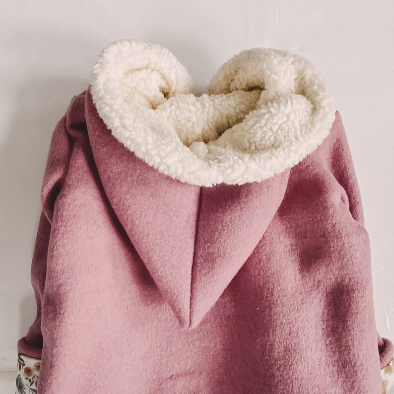 May include: A pink hooded jacket with a white sherpa lining. The hood is folded down and the jacket is made of a soft, fuzzy fabric.