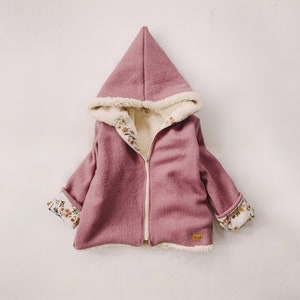 Może przedstawiać: Kurtka z kapturem dla dziecka, wykonana z miękkiej, różowej tkaniny z białym podszewka sherpa. Kurtka ma zamek błyskawiczny i kwiatowy nadruk na mankietach.