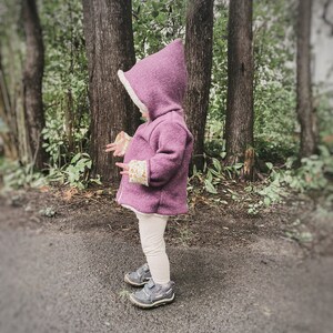 May include: A young child wearing a purple hooded jacket with a white fleece lining. The jacket has a floral pattern on the cuffs. The child is standing in a forest setting.