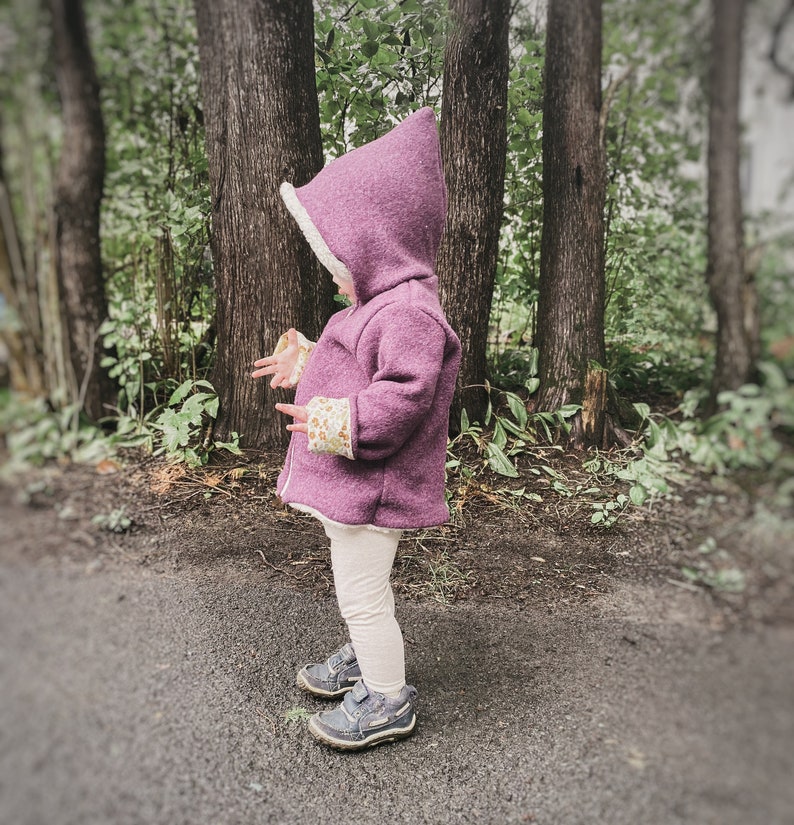 May include: A young child wearing a purple hooded jacket with a white fleece lining. The jacket has a floral pattern on the cuffs. The child is standing in a forest setting.