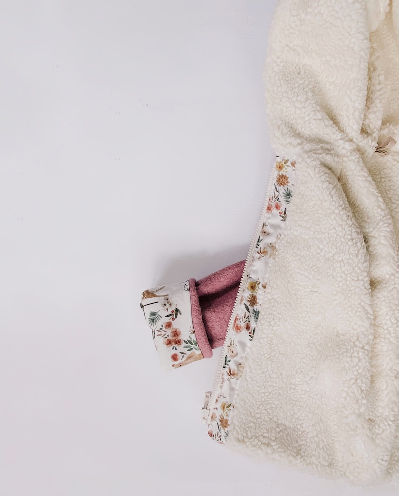 May include: A white fleece jacket with a floral print lining. The lining is visible where the zipper is partially open. The jacket is folded in half and a pink sleeve is tucked inside.