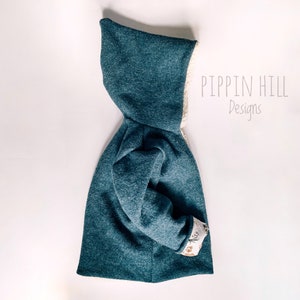 May include: A teal blue hooded jacket with a white sherpa lining. The jacket has a floral print on the cuffs. The text "Pippin Hill Designs" is printed on the right side of the jacket.