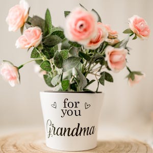 May include: White ceramic planter with pink artificial flowers. The planter has the text "for you Grandma" written on it.