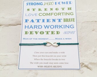 May include: A white card with blue and green text that reads "Strong Hero Power, Strength, Love Comforting, Patient Brave, Hard Working, Devoted Inspire, Man of the Moment...Make a Wish". A teal string bracelet with a silver infinity charm is attached to the card.