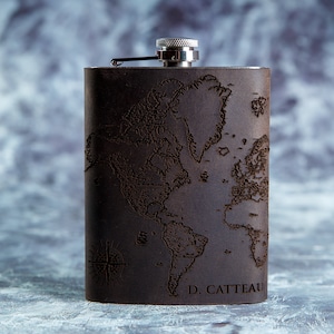 May include: A dark brown flask with a world map design etched into the surface. It features a silver cap and hinged closure. The name "D. CATTEAU" is printed at the bottom. The flask is approximately 13 cm tall.