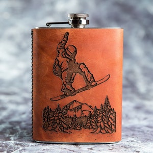 Puede incluir: Una petaca envuelta en cuero marrón con una tapa plateada. La petaca presenta un diseño grabado de un snowboarder en un entorno de montaña.