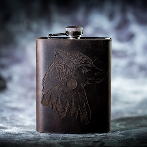 May include: A brown leather flask with a silver top and an engraved design of a wolf's head with a full moon and trees in the background.