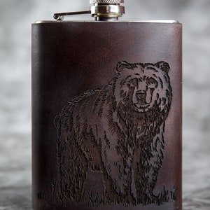May include: A brown leather-covered flask with a silver top and a laser-etched image of a bear.