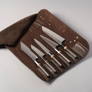 以下が含まれることがあります： 6 本のナイフ用の茶色の革製のナイフロール。ロールが開いており、ナイフが見えます。ナイフは木製のハンドルで、金属製です。