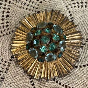 May include: A gold-toned brooch with a cluster of green rhinestones in the center. The brooch has a sunburst design around the rhinestones.