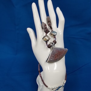 May include: A silver necklace with a large, triangular, pink and brown agate pendant. The necklace is made with small, dark red beads and silver accents.