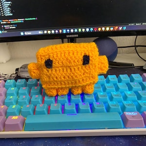 May include: A small, rectangular, bright yellow crocheted plush toy with two dark blue square eyes, two side arms, and two small legs, resting on a mechanical keyboard with light blue and purple keys that feature backlit illumination.