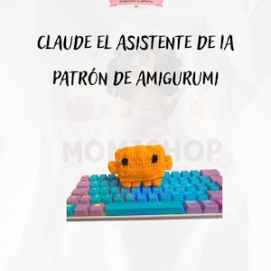 May include: A small orange crocheted amigurumi figure with two black eyes resting on a colorful computer keyboard. The image includes the text MOMISHOP, CLAUDE EL ASISTENTE DE IA, and PATRÓN DE AMIGURUMI, along with illustrations of yarn balls.