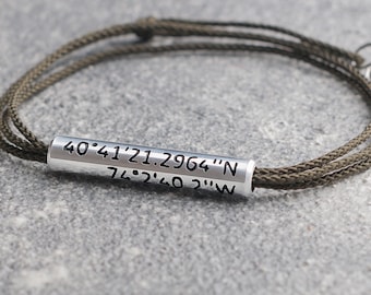 May include: A green cord bracelet with a silver metal bead engraved with the coordinates "40°41'21.2964"N 74°2'40.2"W".