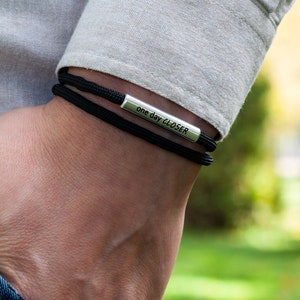 Könnte beinhalten: Ein schwarzes Paracord-Armband mit einem silbernen Barren-Anhänger, auf dem "one day closer" steht.