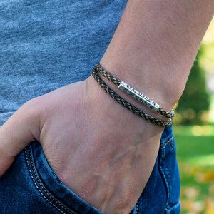May include: A double-layered bracelet with a silver charm engraved with coordinates. The bracelet is made of a dark green and brown cord.