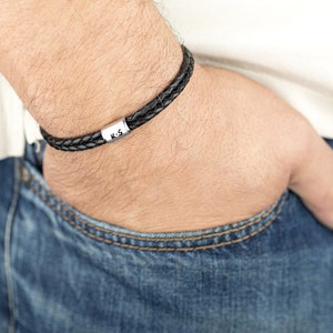 Op de afbeelding: Een zwart leren armband met een zilveren bedel met de tekst "K-S".