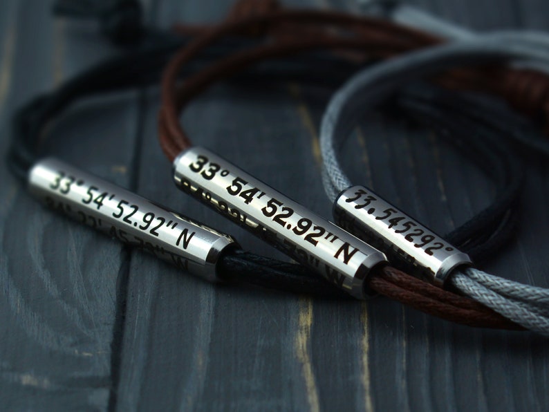 May include: Three leather bracelets with silver metal beads engraved with coordinates. The bracelets are in black, brown, and gray.