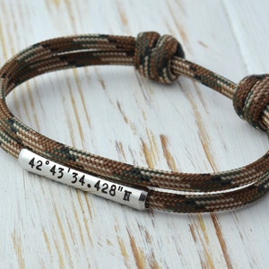 May include: A brown and green camouflage paracord bracelet with a silver metal tag engraved with the coordinates "42&deg; 43' 34.428" N"