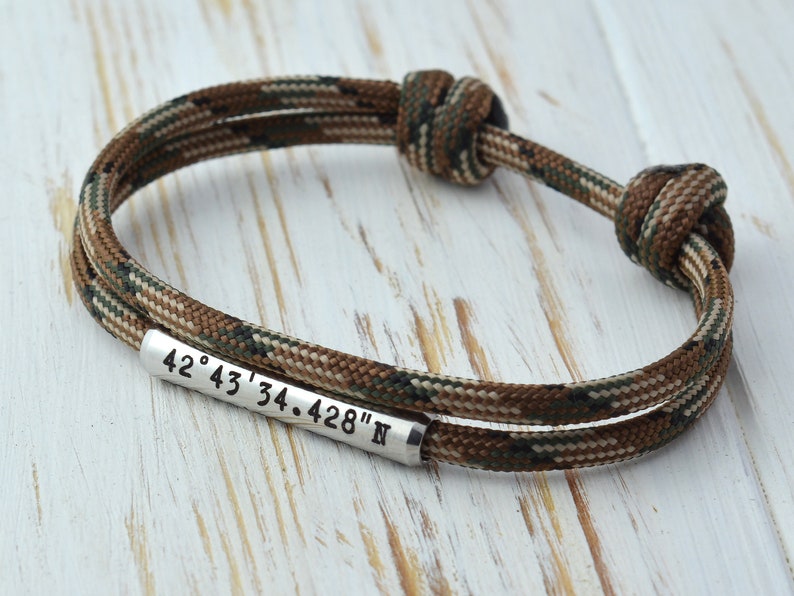 May include: A brown and green camouflage paracord bracelet with a silver metal tag engraved with the coordinates "42&deg; 43' 34.428" N"