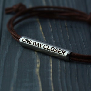May include: A brown leather bracelet with a silver metal charm that reads "ONE DAY CLOSER".