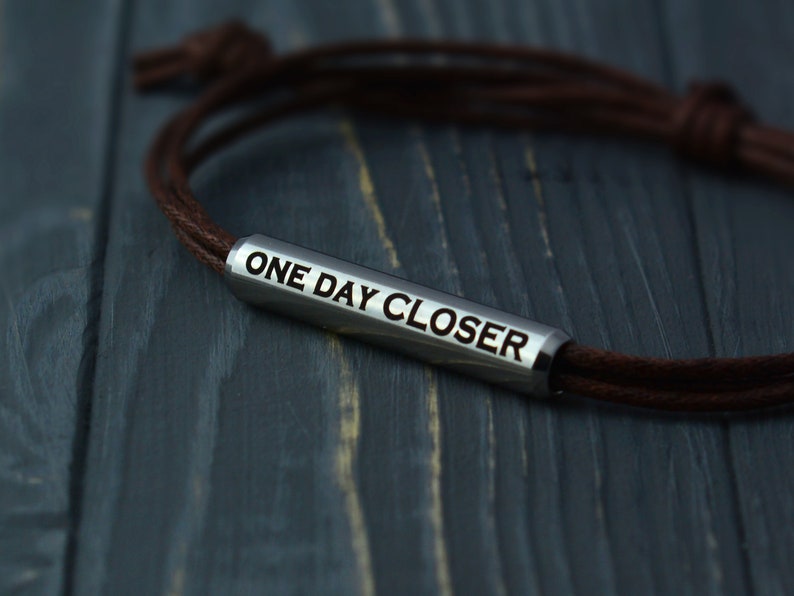 May include: A brown leather bracelet with a silver metal charm that reads "ONE DAY CLOSER".